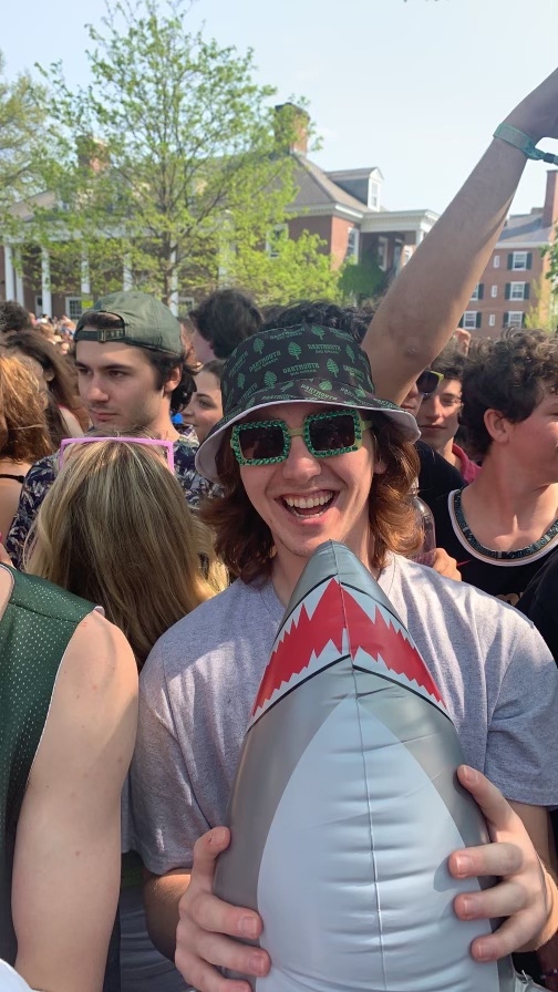 (Me) Garrett Crouch holding a inflatable shark toy, wearing a green Dartmouth bucket hat and green beaded sunglasses smiling in a dense crowd of people on frat row on Dartmouth's campus 