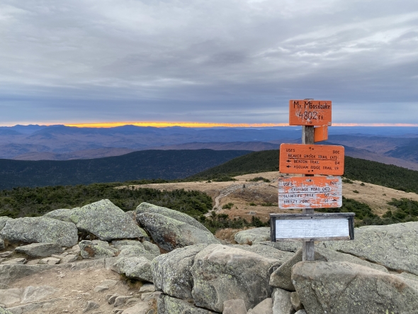 A view of the summit of Mount Moosilauke with a cloudy sky and glowing orange horizon.