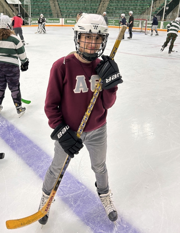 a Dartmouth student (the author of this post) in a maroon crewneck with the letters A and theta on it, gray jeans, hockey skates, and a helmet, holding a hockey stick on the ice