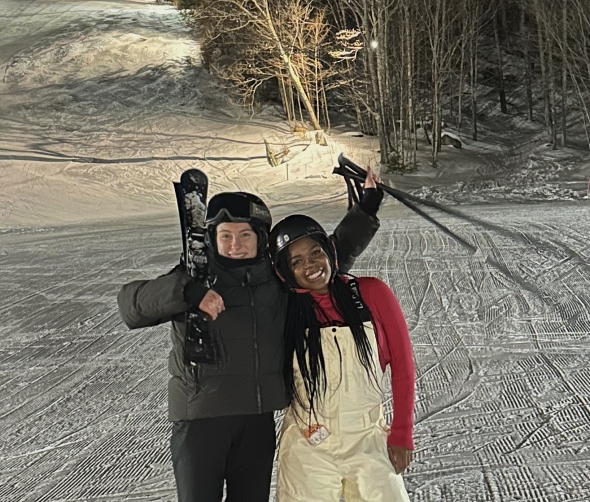 Two smiling skiiers in ski equipment, ski poles in hand, smiling broadly in front of a snowy hill. 