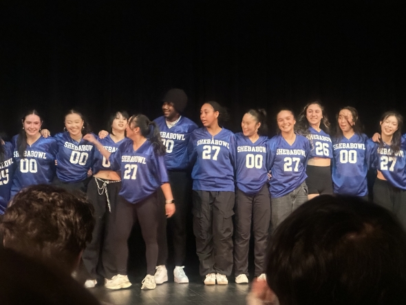 A dance group is smiling as they stand in a line together, wearing blue jerseys with respective numbers on the front.