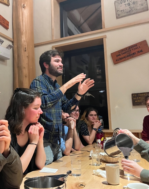 Kevin, a white man wearing a plaid shirt, is standing up from a long table. Many people look to him from their seats.