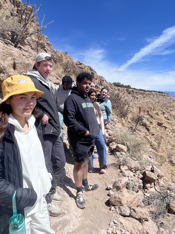 Some of the Dartmouth students standing on the side of the Puye cliffs with the desert cliff landscape surrounding. Bright blue sky in the background.