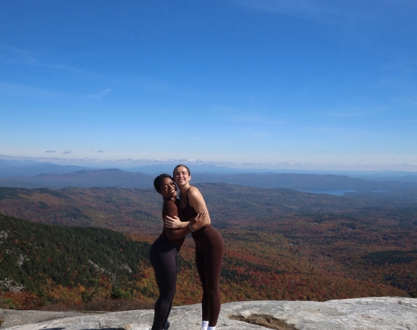 Two girls are smiling and facing the camera while embraced into a hug, posing in front of a scenic landscape that the mountain overlooks, displaying vibrant views of fall foliage and colorful trees beyond the rocky terrain.