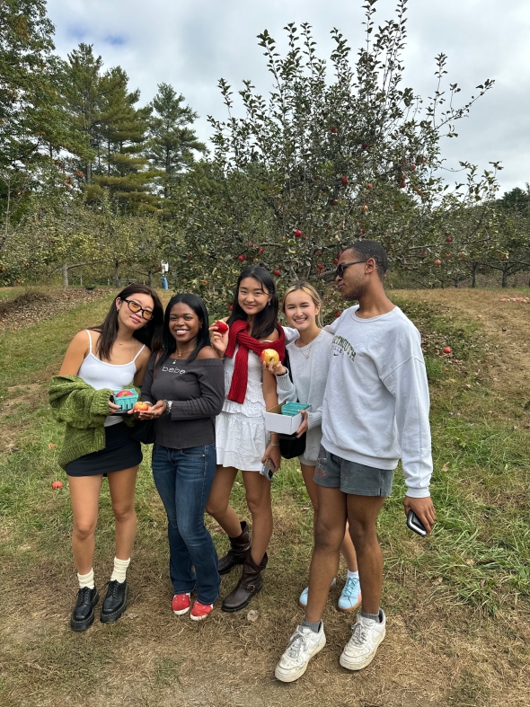 Five people smile brightly while standing in the middle of an apple-picking farm, holding apples and containers filled with fruit. The vibrant farm landscape surrounds them, adding to the cheerful atmosphere of the moment.