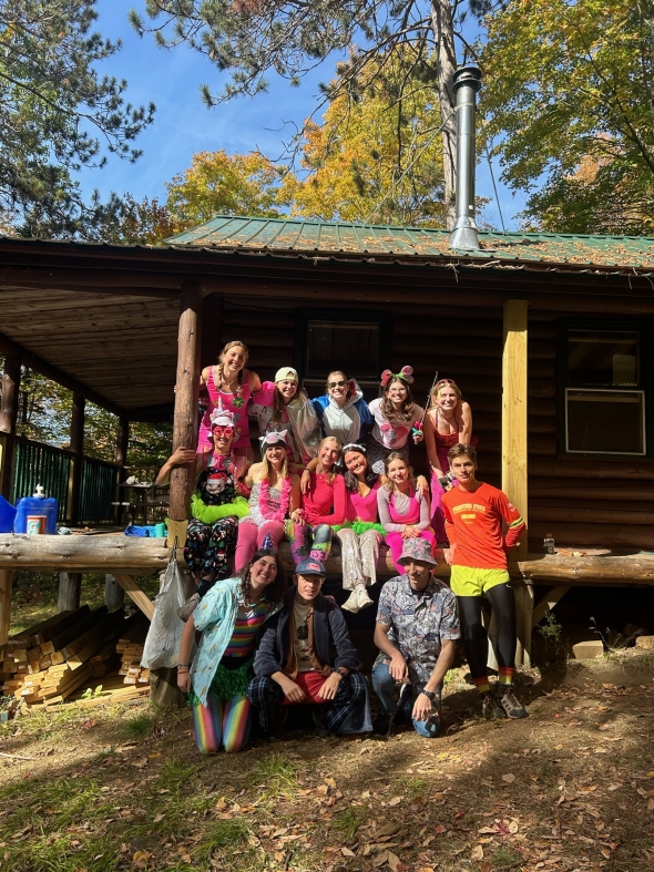 A group of Dartmouth students dressed in flair pose for a group photo in front of a log cabin.