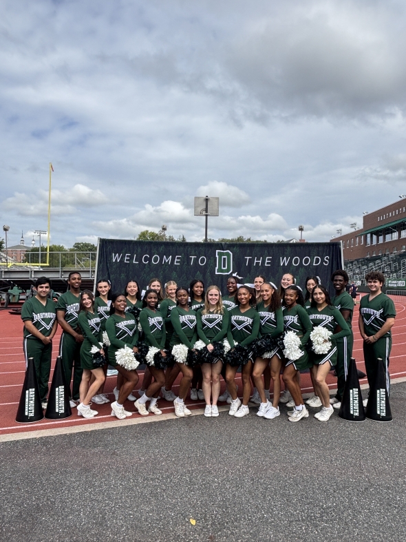 A cheer team poses in front of a Dartmouth banner that reads 'Welcome to the Woods.' Four small megaphones are placed on the ground at the sides of the cheerleaders, framing the group as they smile proudly in their team formation.