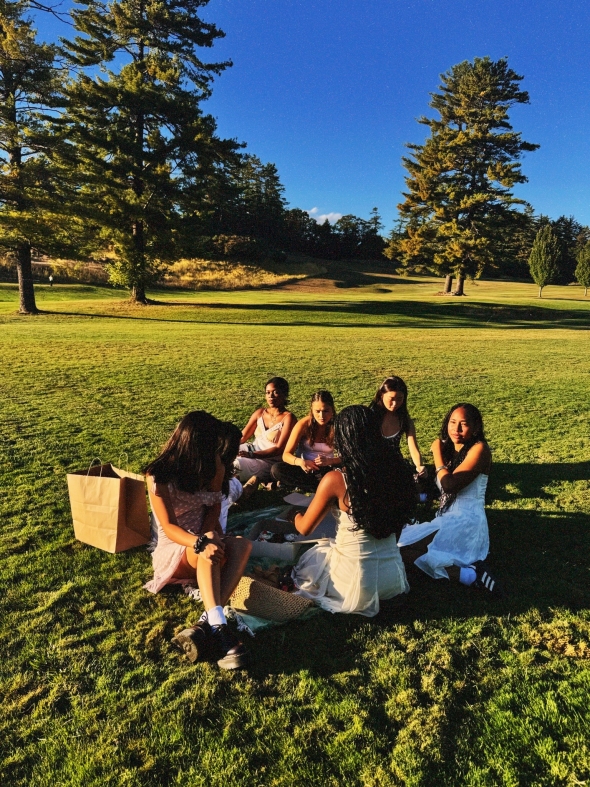 A group of girls enjoy a picnic in the middle of a grassy field, surrounded by scattered trees in the distance. They sit together in the peaceful, open landscape.