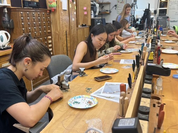 A row of students sit at a wood workbench making jewelry.