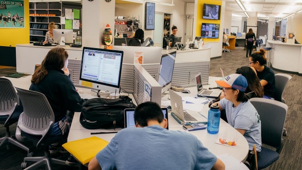 Students sit at desks looking at monitors in a well-lit library space.
