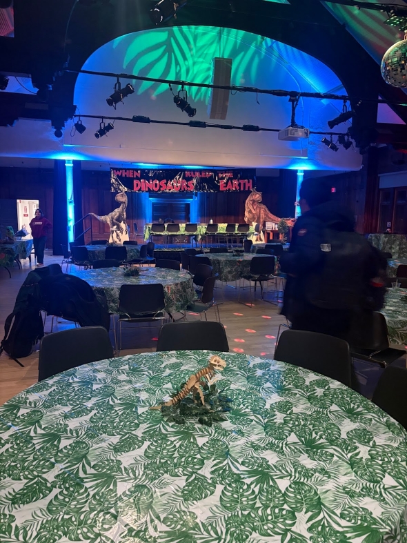 A dark room with chairs and tables set up. The tablecloths are jungle leaf-shaped, and there are cutouts of dinosaurs in the background.