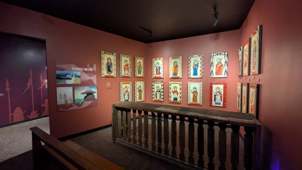  A room in the Indian Pueblo Cultural Center museum with the walls painted a rusty red color and a sign that says 'Religious Friction' with Christian saints surrounded by Native American motifs. 