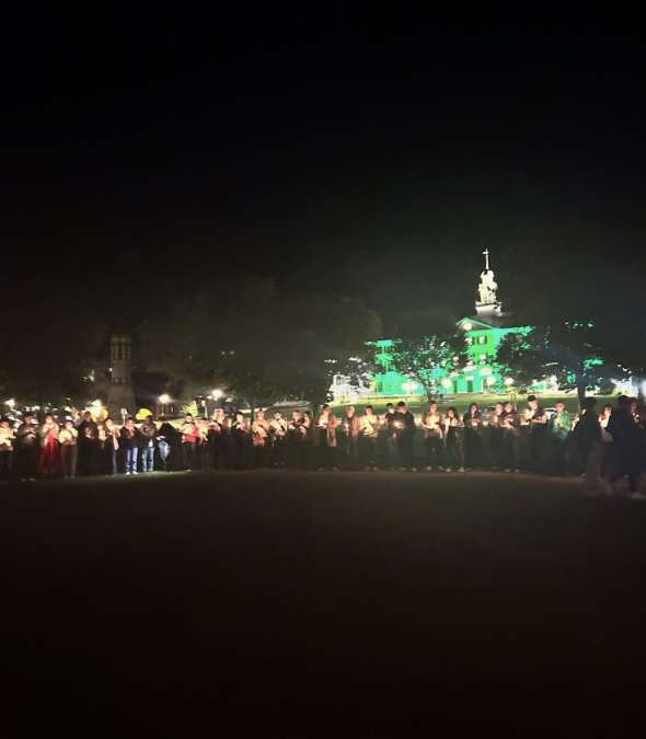 people are standing in a semi-circle in front of a building covered in green light. They are holding candles.