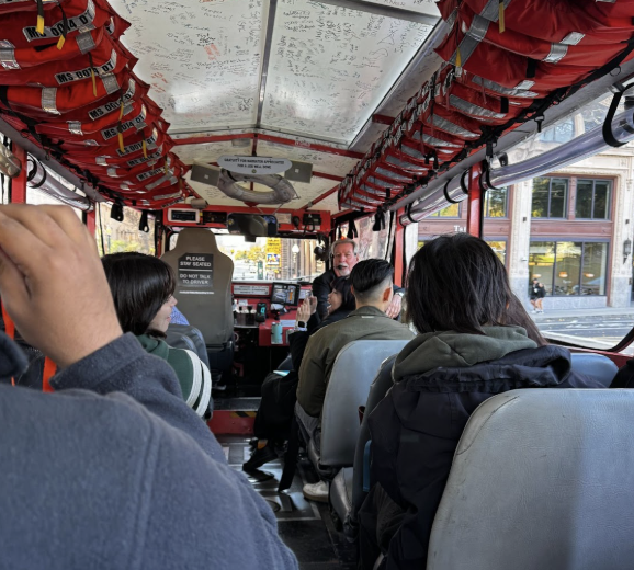 Image featuring the inside of the quack-mobile. The image features the inside of a vehicle with several seats with people sitting on them. A tour guide can be seen speaking in the background.