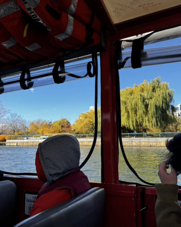 Picture featuring the inside of the quack-mobile on the water. Several seats and people sitting on them can be seen. Outside the window, one can clearly see the vehicle gliding through one of Boston's rivers.
