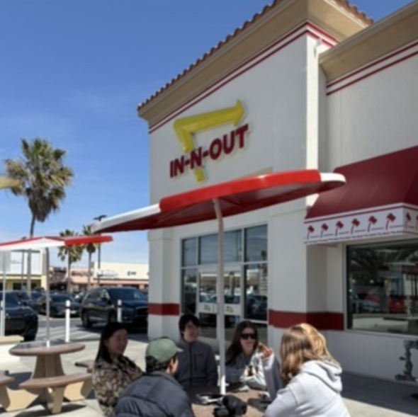 A group of people sitting under a shaded outdoor booth in front of a fast food restaurant. The restaurant has a logo with the word "IN-N-OUT"