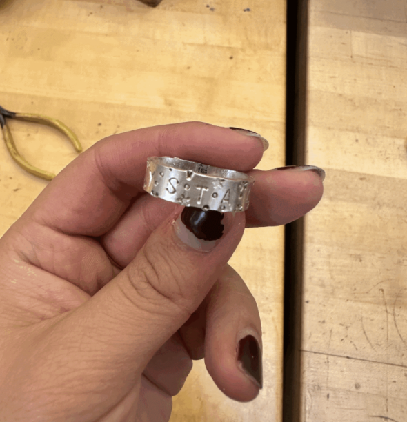 An image of me, Crystal, holding a silver ring with the letters "STA" engraved on it. The background is of a wooden table.