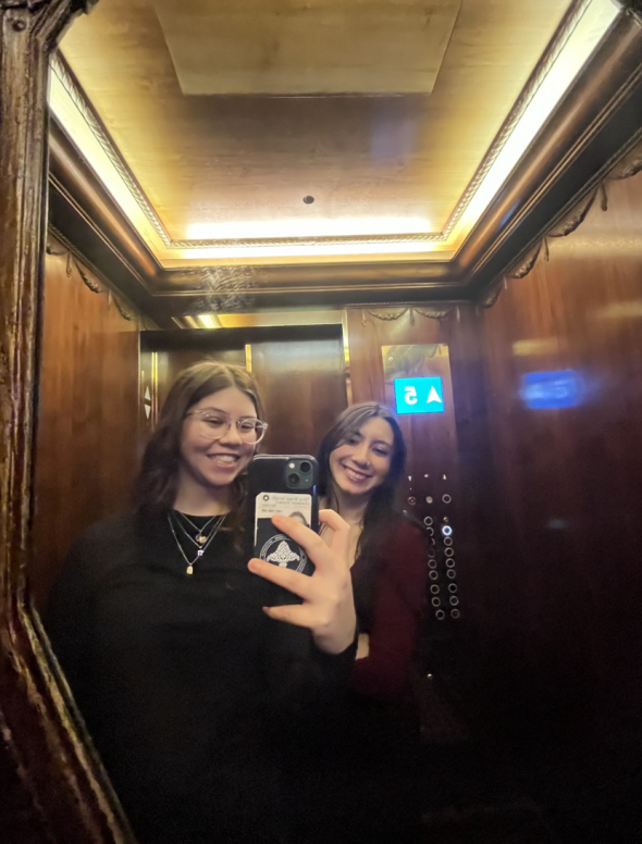 A mirror selfie with two women smiling and looking at the camera. They are in an elevator with wood paneling
