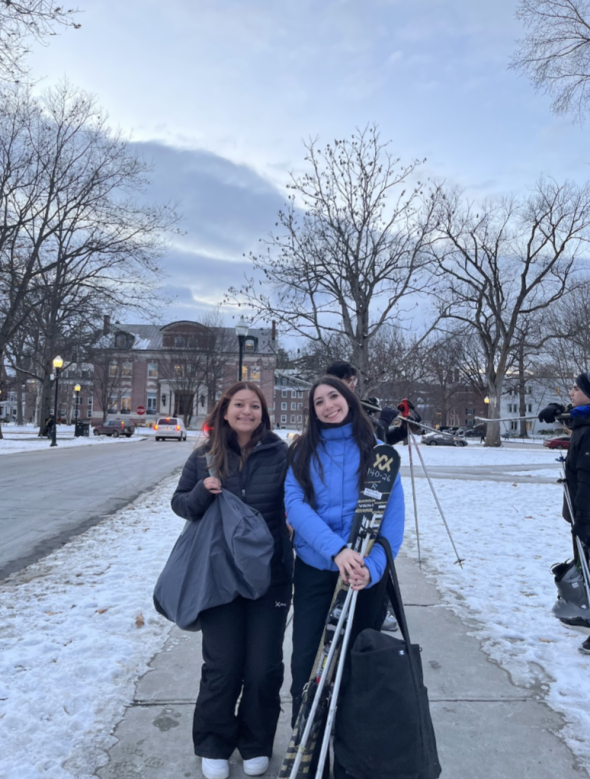 two women hold skis and smile at the camera. The sidewalk is snowy and the blue sky is dotted with clouds.