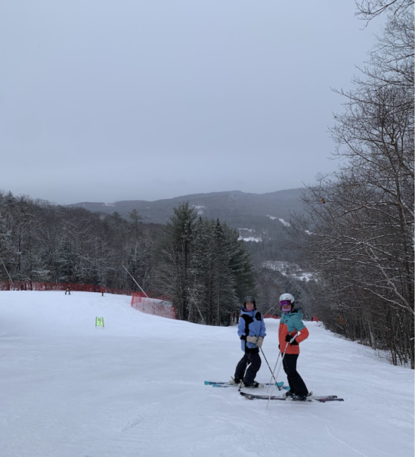 two women pose on skies and look up the mountain. The snowy ground slopes downward behind them.