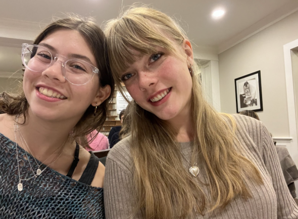 Serena and her friend Grace lean their heads together and smile. Serena is on the left with a blue sweater and Grace is on the right with a beige sweater. Tan walls are behind them.