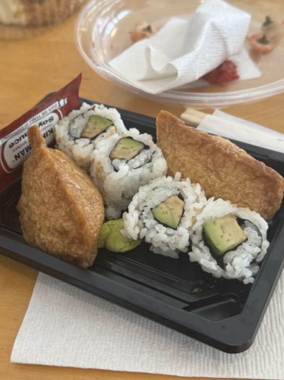 A plastic container with avocado sushi rolls and inari (sweet tofu pockets) sits on a table, accompanied by a soy sauce packet.