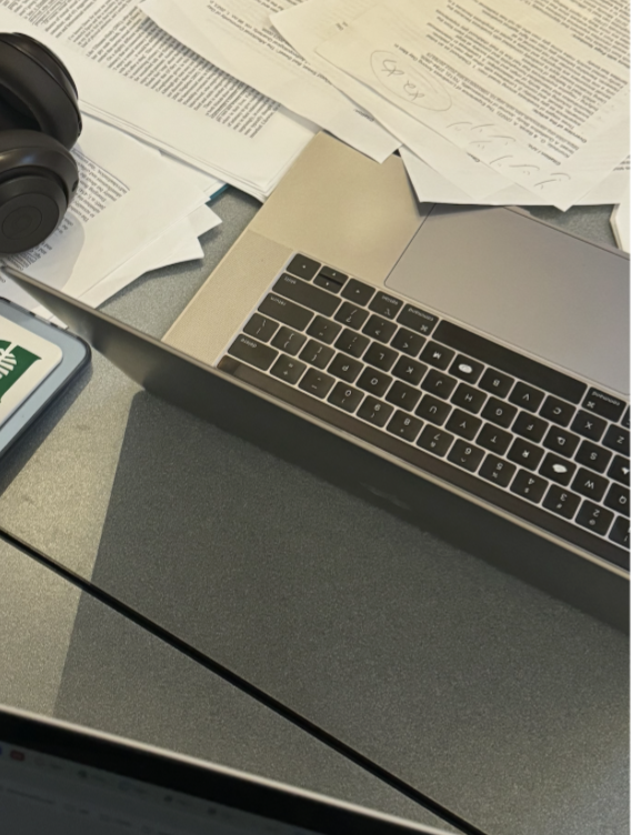 A close-up of a study desk with an open laptop, scattered papers with handwritten notes, and a pair of black headphones resting nearby. A Dartmouth sticker is visible on the desk.