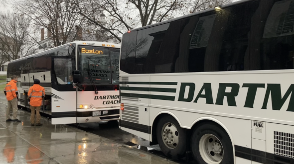 Two Dartmouth Coaches to Boston lined up at the bus stop. Two people in orange vests stand next to the bus further from the camera.