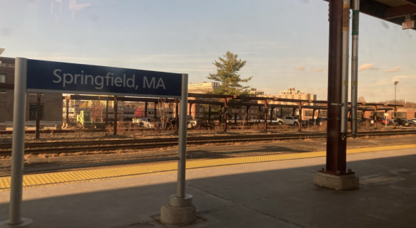 An image of the Springfield, Massachusetts Amtrak station. The station is a flat concrete platform covered by a roof, to the left of the image is a blue sign which reads "Springfield, MA", behind the platform are railroad tracks.