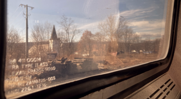 A view out of the train window. The sky is blue with sparse clouds, the landscape is dotted with bare trees, buildings, and piles of wood.