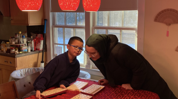 A young boy holding a box of mahjong tiles speaks to Iman over a table. In the background is a window, a kitchen to the right, and decorative fans to the left.