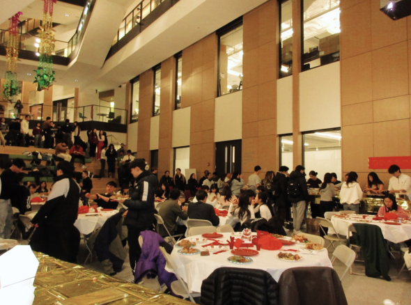 A view of the lunar new year banquet. People line up along the wall, waiting to be served food. In the foreground are people sitting around multiple circular tables, enjoying their food. 
