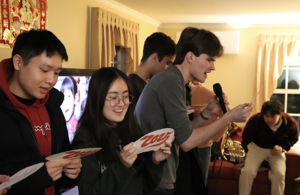 Four people sing while standing in a line in front of a television, each holding a piece of paper with lyrics. The third person is also holding a microphone.