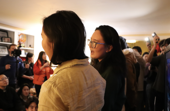 A view of the back of two singers. An audience sits or stands in the background, watching the performance and waving their camera flashlights.