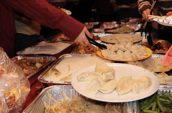 Four yellow and white plates of dumplings balance on top of a table full of aluminum catering trays of various Chinese dishes. Someone reaches for a dumpling with plastic tongs.