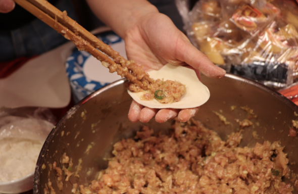 A view of dumpling making. Someone's hand holds a dumpling wrapper, and a pair of chopsticks to add dumpling filling in the other. In the background is a bowl of dumpling filling.