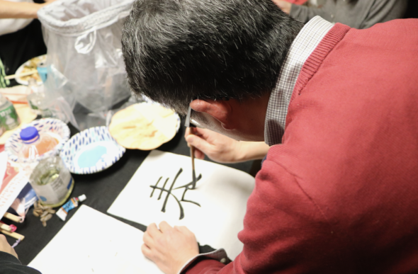A view over-the shoulder of a person wearing a red sweater. They are using a brush to write a Chinese character in black ink. In the background are bowls and bottles of other colors.