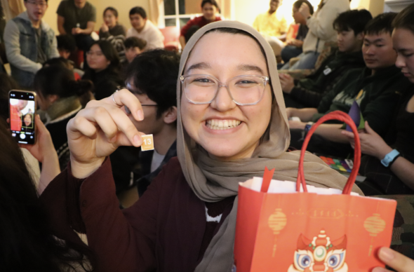 A smiling person holds up a piece of paper with the numbers "13" on it in one hand. With the other hand, they hold a red paper bag decorated with Chinese New Year graphics. People are seated in the background.