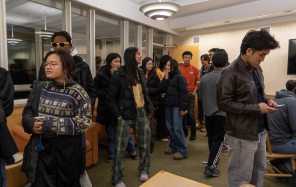 People stand and chat in a line to the left side of the image. On the right side of the image, a few people are seated.