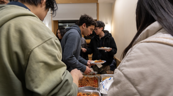 A view of a table full of food, overlooking two people's shoulders. The food is held in aluminum serving trays. There is a person serving themselves food in the middle of the image.
