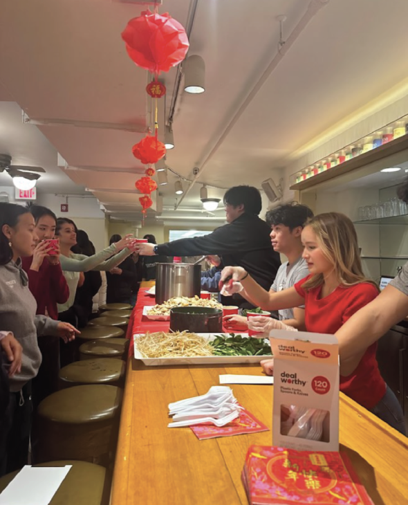 Students serve and receive cups of phở. Three students stand behind the bar of One Wheelock. On the bar are plates of toppings, noodles, and pots of broth. More students line up on the other side of the bar waiting for a cup. Red decorations are hung abov