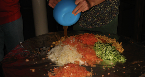 A view of the yee sang before tossing. Salmon, wonton crackers, pomelo pieces, and shredded carrots, white radish, and cucumber are piled on a table covered with aluminum-foil. Someone is pouring sauce out of a blue bowl on top.