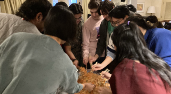A view of the lou sang. Multiple students crowd around the aluminum foil-covered table, tossing the salad with wooden chopsticks. Four students have their backs facing the camera.