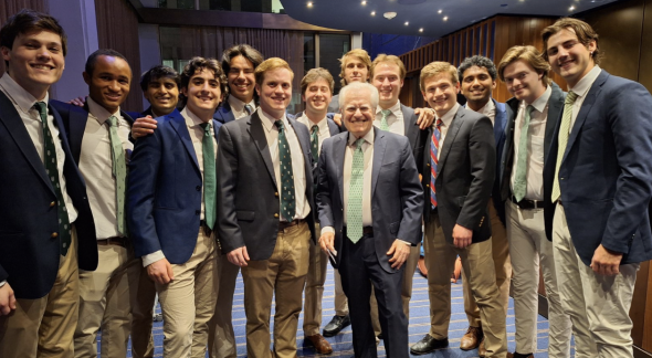 An image of a large group of students in blue suits meeting an alumni. The students appear to be in a performance hall.
