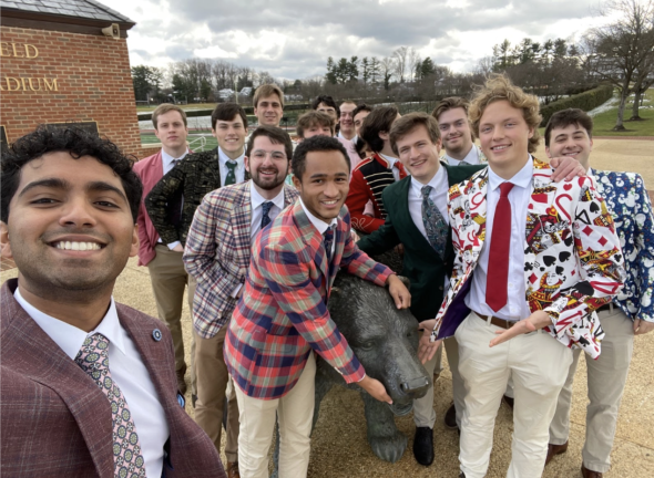 A group of students in colorful suits outside. They are holding a metal bear statue. The background is cloudy and the students are next to a stadium.