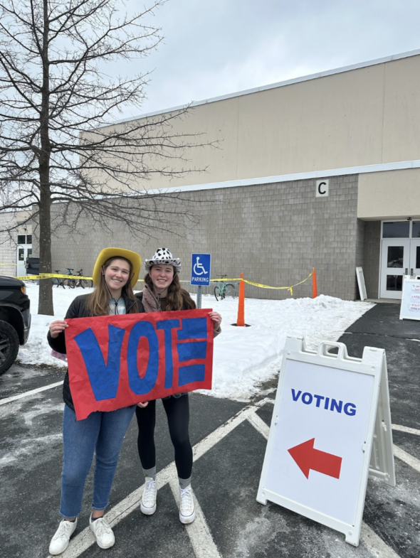 Eleanor and Bea holding a sign that says "VOTE" in front of Hanover High School. They are both smiling and wearing silly cowboy hats.