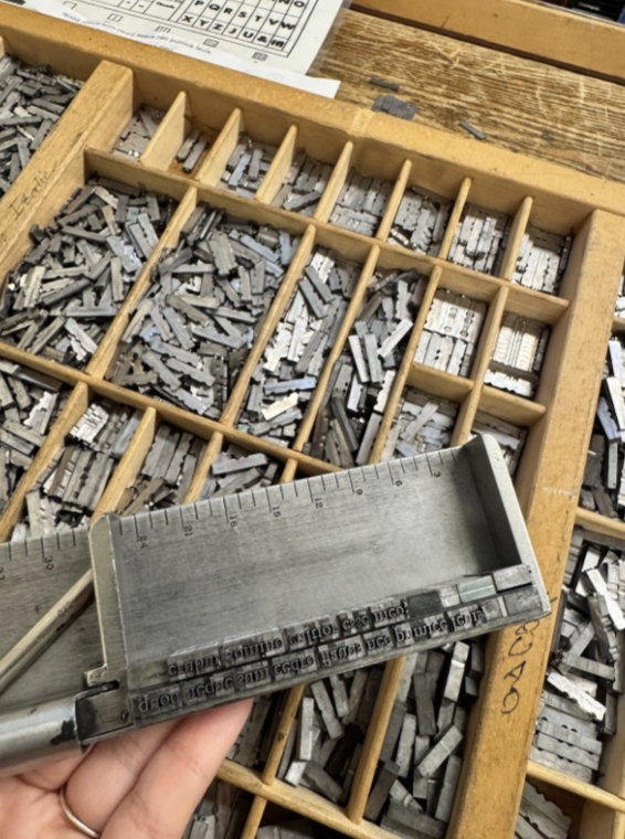 A job stick containing letters and spacers held above a case of letters and spacers.