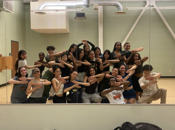 A group of students posing in a group. The photo is taken as a mirror reflection. The students are in a practice space and are holding their hands in an interlocked position.