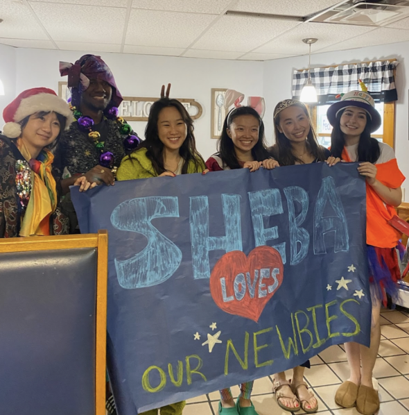 A group of students inside a restaurant, wearing various articles of clothing and holding a paper sign that says "Sheba loves our newbies"