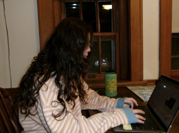 Serena sits at the table with her hands on her computer in the midst of typing. Windows are behind her and her green water bottle rests on the side of the table that is closer to the windows. 